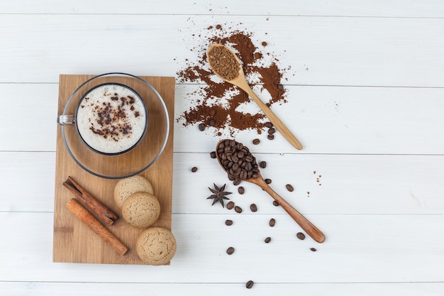 Wat koffie met gemalen koffie, koffiebonen, kaneelstokjes, koekjes in een kopje op houten en snijplank achtergrond, plat leggen.