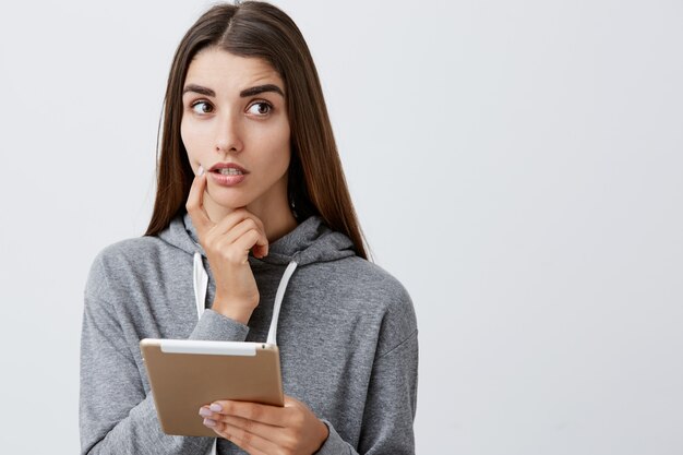 Wat zal ik kopen. Sluit omhoog van charmant knap jong Kaukasisch meisje met donker lang haar in toevallige grijze hoodie die vinger dichtbij lippen houden, kijkend door kleding voor prom