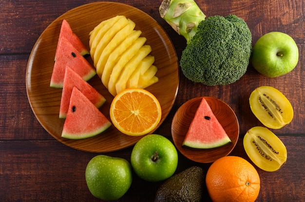 Watermeloen, sinaasappel, ananas, kiwi in plakjes gesneden met appels en broccoli op een houten plaat en houten tafel.