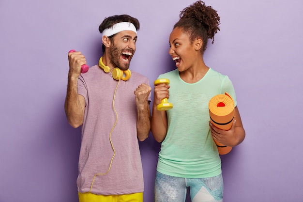 Wees sterk en gezond. Emotionele diverse vrouw en man schreeuwen van vreugde, kijken elkaar aan, staan met sportuitrusting, trainen spieren met kleine halters, dragen fitnessmat, trainen in de sportschool