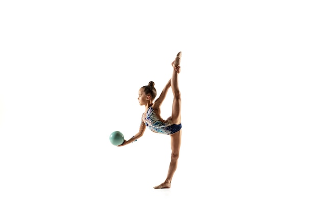 Weinig flexibel meisje dat op witte muur wordt geïsoleerd. Weinig vrouwelijk model als ritmische gymnastiekkunstenaar in heldere maillot. Gratie in beweging, actie en sport. Oefeningen doen met de bal.