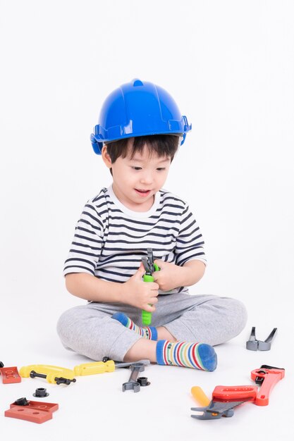 Weinig jongen die blauwe helmzitting draagt en met het stuk speelgoed van het bouwmateriaal speelt op wit