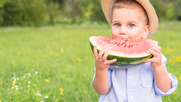 Gratis foto weinig jongen die grote rode plak van watermeloen in het park eet