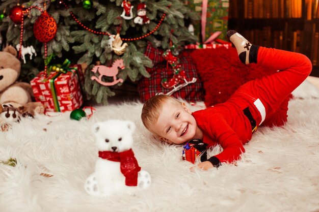 Weinig jongen in feestelijke rode kostuumspelen vóór een Kerstboom