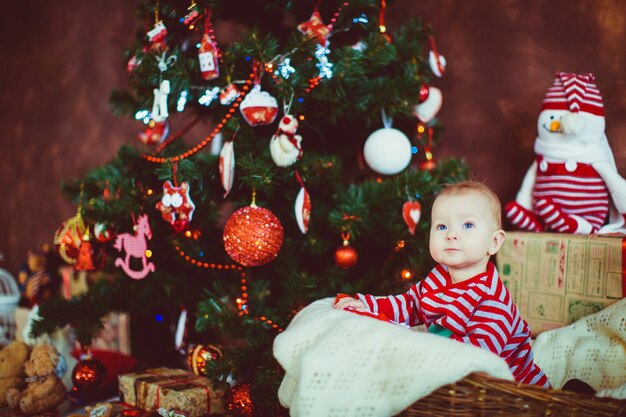 Weinig jongen in gestripte pyjama zit vóór een Kerstboom