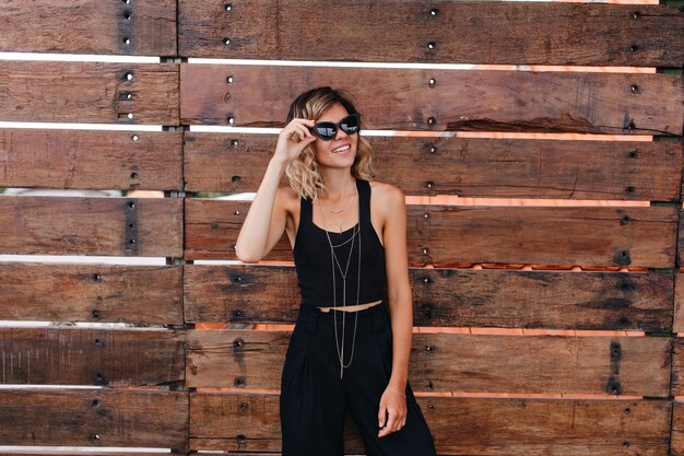 Welgevormde jonge vrouw in zwarte broek met plezier tijdens fotoshoot. aangenaam krullend vrouwelijk model draagt een zonnebril en een stijlvolle tanktop.