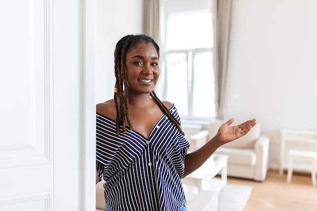 Welkom portret van een vrolijke afrikaanse vrouw die een bezoeker uitnodigt om zijn huis binnen te gaan, een gelukkige jonge vrouw die in de deuropening van een modern appartement staat en een woonkamer met de hand toont