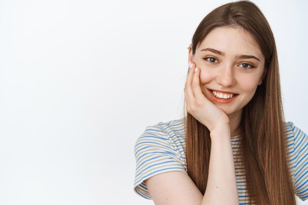 Welzijn van vrouwen. Schattig meisje raakt haar schone gezicht aan met een gloeiende huid, natuurlijke uitstraling, glimlachend gelukkig op camera, witte achtergrond