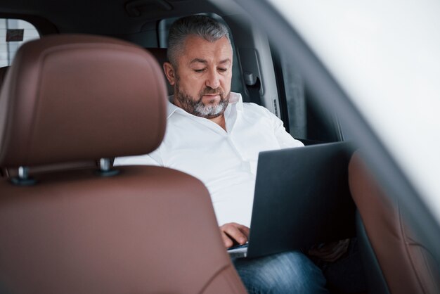 Werken aan een achterkant van de auto met behulp van zilverkleurige laptop. Senior zakenman