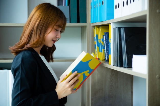 Werkende vrouw die een boek van planken in bibliotheek leest