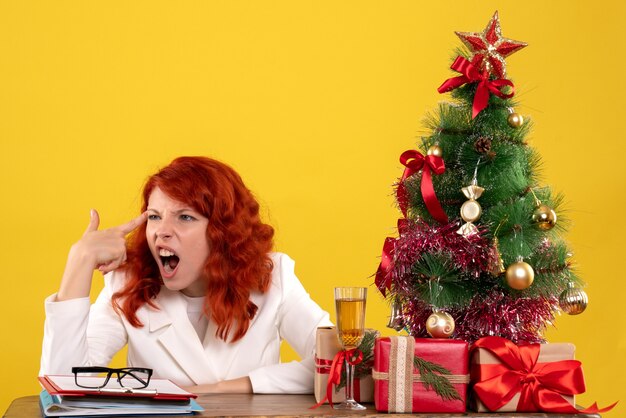 werkneemster zittend achter tafel met kerstcadeautjes en boom op geel