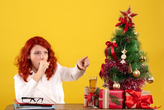 werkneemster zittend achter tafel met kerstcadeautjes en boom op geel