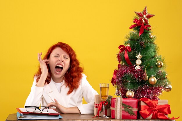 werkneemster zittend achter tafel met kerstcadeautjes en boom op geel