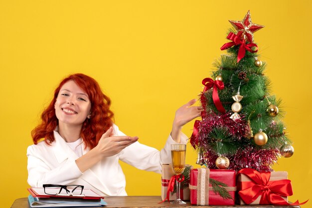 werkneemster zittend achter tafel met kerstcadeautjes en boom op geel