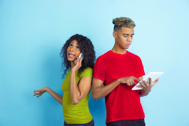 Werkt op tablet, praat via telefoon. jonge emotionele afro-amerikaanse man en vrouw in kleurrijke kleding op blauwe achtergrond. mooi paar. concept van menselijke emoties, gezichtsuitdrukkingen, relaties, advertentie.
