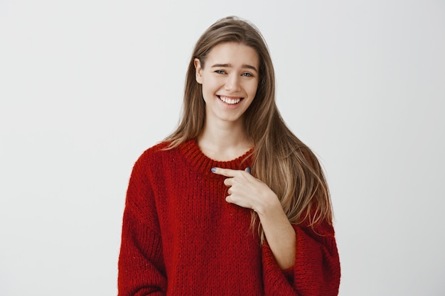 Wie ik, maak me niet aan het lachen. Portret van minachtende zorgeloze emotionele Europese vrouwen in rode losse trui, wijzend naar zichzelf met wijsvinger, lachend en starend met minachting over grijze muur