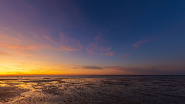 Wijd geschoten van natte strandkust onder een blauwe en gele hemel bij zonsondergang