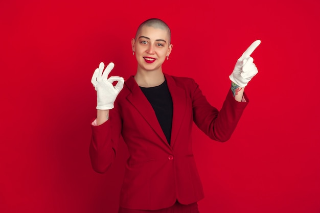 Wijzen, tonen. Portret van jonge blanke kale vrouw geïsoleerd op rode muur. Mooi vrouwelijk model in jasje. Menselijke emoties, gezichtsuitdrukking, verkoop, advertentieconcept. Gekke cultuur.