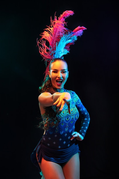 Wijzend. Mooie jonge vrouw in Carnaval, stijlvol maskeradekostuum met veren op zwarte muur in neonlicht. Copyspace voor advertentie. Feestdagen, dansen, mode. Feestelijke tijd, feest.