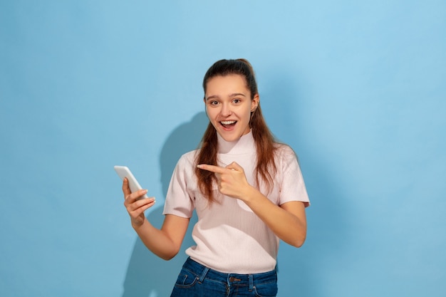 Wijzend op smartphone, glimlachend. Het portret van het Kaukasische tienermeisje op blauwe achtergrond. Prachtig model in vrijetijdskleding. Concept van menselijke emoties, gezichtsuitdrukking, verkoop, advertentie. Copyspace. Ziet er blij uit.