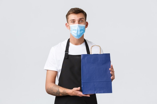 Winkel, winkelen tijdens covid-19 en concept voor sociale afstand. Vriendelijke beleefde verkoper, barista met medisch masker en zwarte schort, zet gekocht artikel in eco-tas, staande witte achtergrond