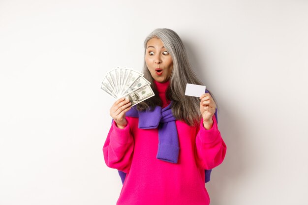 Winkelconcept. Gelukkige aziatische senior vrouw die verbaasd naar geld kijkt en plastic creditcard toont, staande op een witte achtergrond.