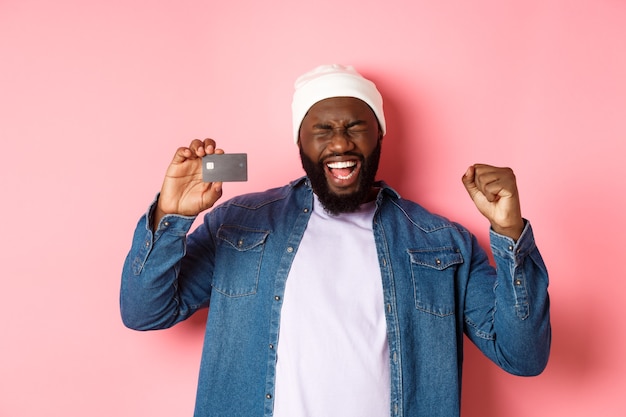 Winkelconcept. Gelukkige zwarte man verheugt zich, schreeuw van vreugde en toont creditcard, staande over roze achtergrond.