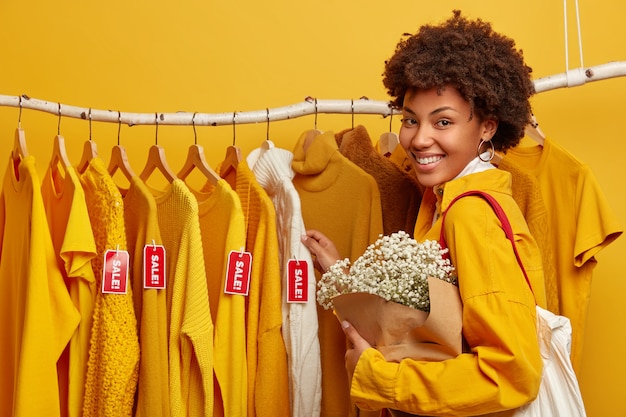 Winkelen levensstijl concept. Positieve vrouwelijke shopper brengt weekend door in modieuze winkel, boeket houdt, staat in de buurt van kledingrek. Grote uitverkoop