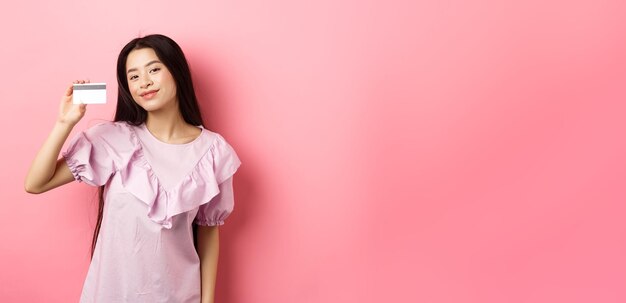 Winkelend jong koreaans meisje in jurk die creditcard toont die tevreden glimlacht naar de camera die op roze staat