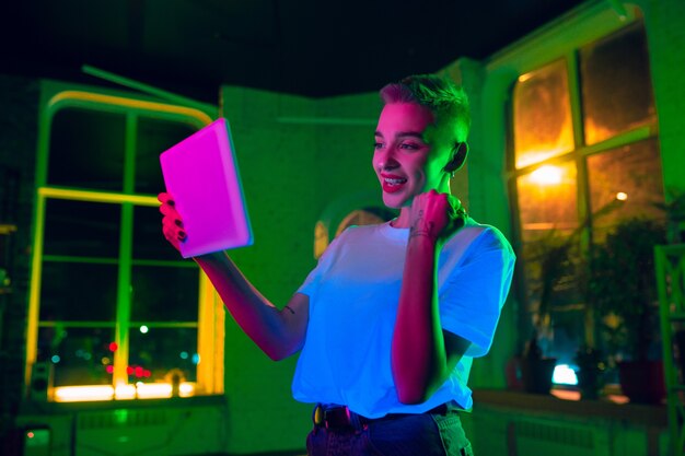 Winnen. Filmisch portret van stijlvolle vrouw in neon verlicht interieur. Afgezwakt als bioscoopeffecten, heldere neon-kleuren. Kaukasisch model met behulp van tablet in kleurrijke lichten binnenshuis. Jeugd Cultuur.