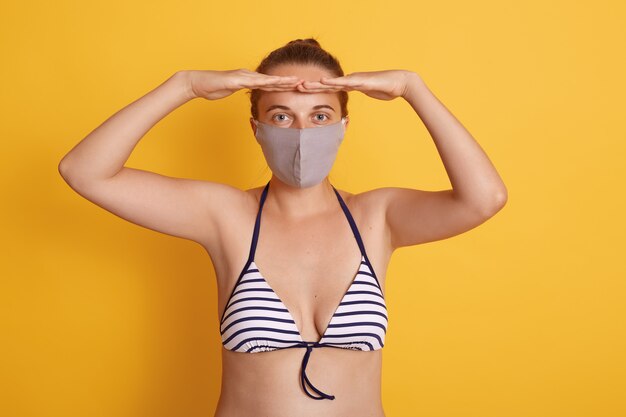 Winsome vrouw in badpak en medisch masker poseert tegen de gele muur, houdt beide handpalmen dichtbij het voorhoofd, kijkt in de verte en draagt een beschermend masker tijdens de quarantaine.