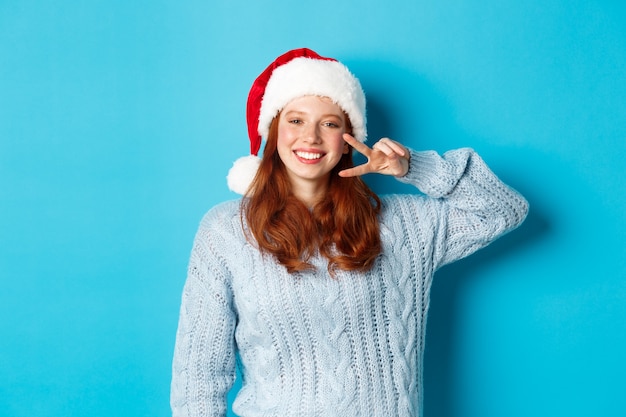 Wintervakantie en kerstavond concept. Gelukkig tienermeisje met rood haar, met kerstmuts, genietend van Nieuwjaar, vredesteken tonen en glimlachen, staande over blauwe achtergrond.