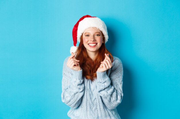 Wintervakantie en kerstavond concept. Hoopvol roodharig meisje in kerstmuts, wensen doen op Kerstmis met gekruiste vingers, kerstmuts dragen, staande over blauwe achtergrond
