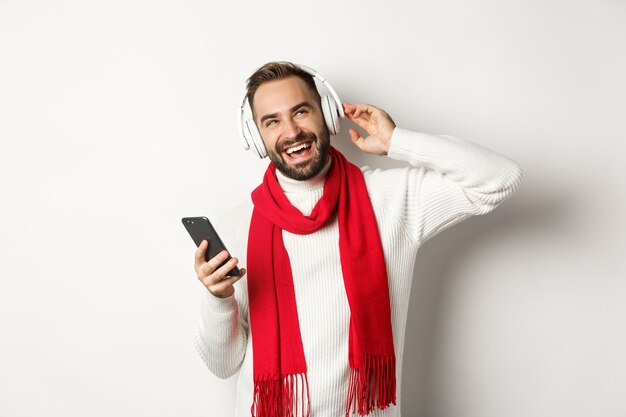 Wintervakantie en technologie concept. Man die geniet van het luisteren van muziek in een koptelefoon, tevreden kijkt, smartphone vasthoudt, trui met sjaal draagt, witte achtergrond