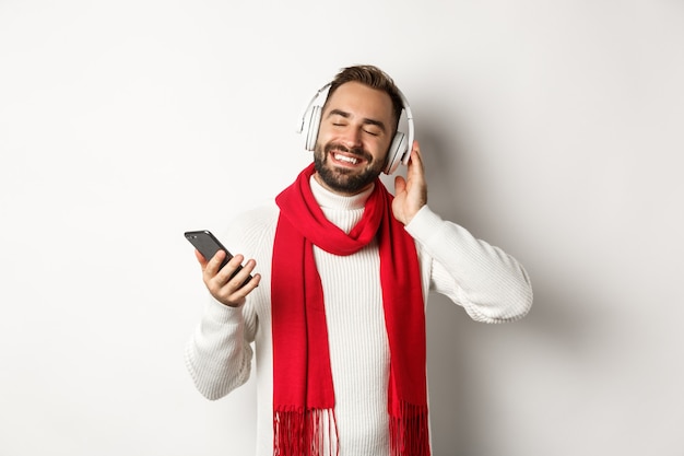 Wintervakantie en technologie concept. Tevreden man die muziek luistert in een koptelefoon met gesloten ogen, glimlachend met plezier, met smartphone, witte achtergrond.