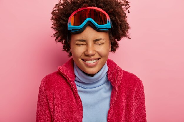 Wintervakantie en vakantie concept. Close-up shot van vrolijke Afro-Amerikaanse vrouw draagt skibril, geniet van vrije tijd, gekleed in coltrui en rode trui, geïsoleerd op roze muur.