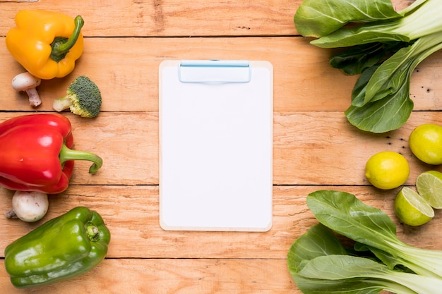 Gratis foto witboek op klembord dichtbij de groene paprika's; broccoli; paddestoel; bokchoy en citroen op houten bureau