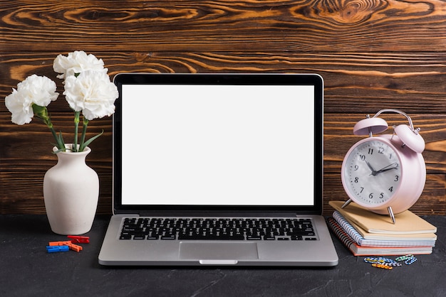 Witte bloemen in de vaas; laptop en wekker op notebooks tegen houten achtergrond