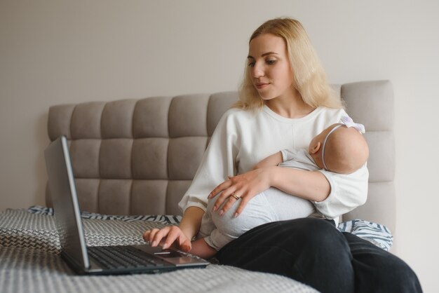 Witte jonge moeder zit met baby op bed en werkt op laptop, werk thuis at