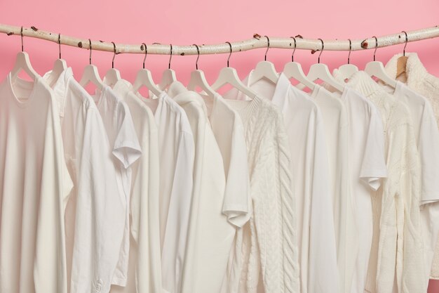 Witte stevige kleren die in één rij op houten rekken hangen tegen roze achtergrond. Grote keuze voor vrouwen in winkel.