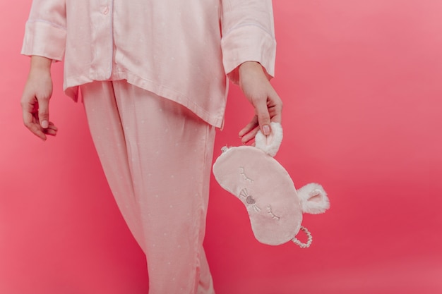 Witte vrouwelijke model poseren in katoenen pyjama's op helder roze muur. Binnenfoto van vrouw draagt nachtkleding met slaapmasker in de hand.