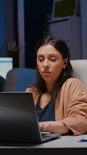 Workaholic ondernemersvrouw die aan een bureautafel zit en financiële afbeeldingen analyseert met behulp van een laptopcomputer