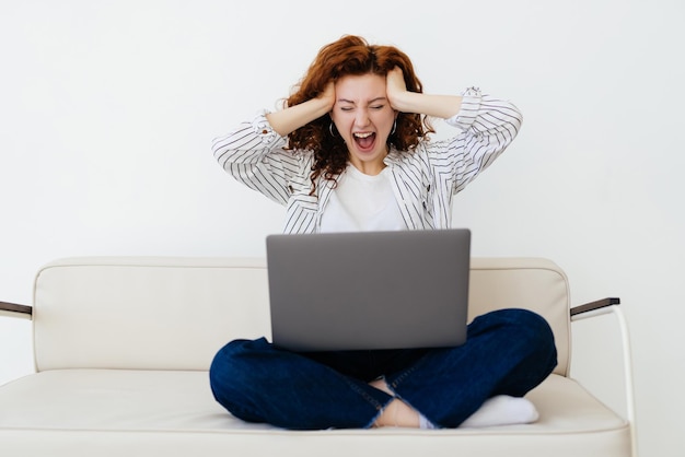 Wow Leuke roodharige vrouw met kunstledemaat zittend op de grijze bank en kijkend naar het laptopscherm terwijl ze geschokte en verraste emoties voelt Tijd thuis doorbrengen concept