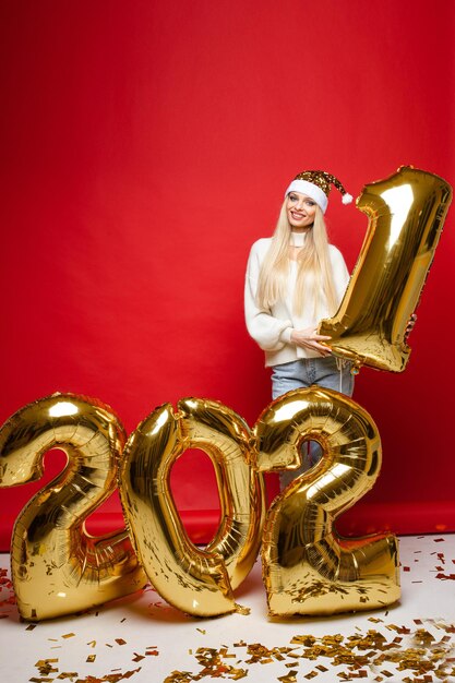 Xmas glimlachend jong meisje in kerstmuts met gouden ballonnen in 2021 vorm op rode achtergrond voor kerstvakantie ontwerp. Hoge kwaliteit foto