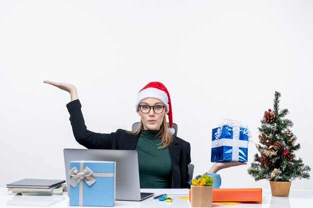 Xmas stemming met verrast jonge vrouw met kerstman hoed en het dragen van een bril zittend aan een tafel met cadeau wijst iets op witte achtergrond