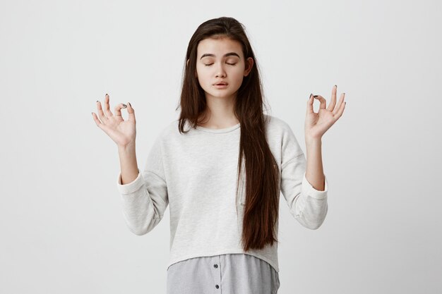 Yoga en meditatie. Mooie terloops geklede donkerbruine vrouw die ogen gesloten houdt terwijl het mediteren, ontspannen, kalm en vreedzaam voelt