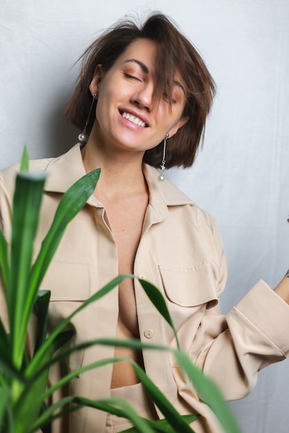 Zacht binnenportret van Kaukasische zachte vrouw die beige kostuum draagt zonder bustehouder, die zich achter grijze palm tropische plant stelt.