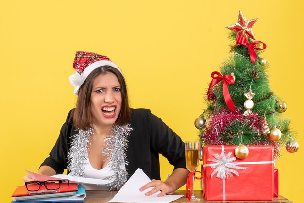 Zakelijke dame in pak met kerstman hoed en nieuwjaarsversieringen boos voelen en zittend aan een tafel met een kerstboom erop op kantoor