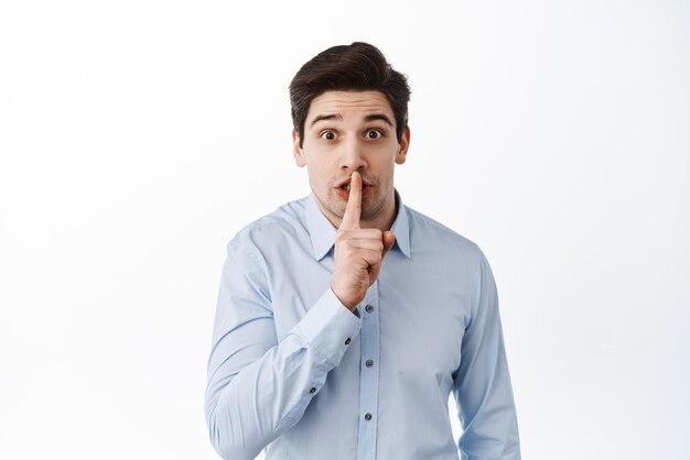 Zakelijke man in hemd die vraagt om stil te blijven roddelen op kantoor terwijl hij zijn vinger tegen de lippen houdt en opgewonden op een witte achtergrond kijkt