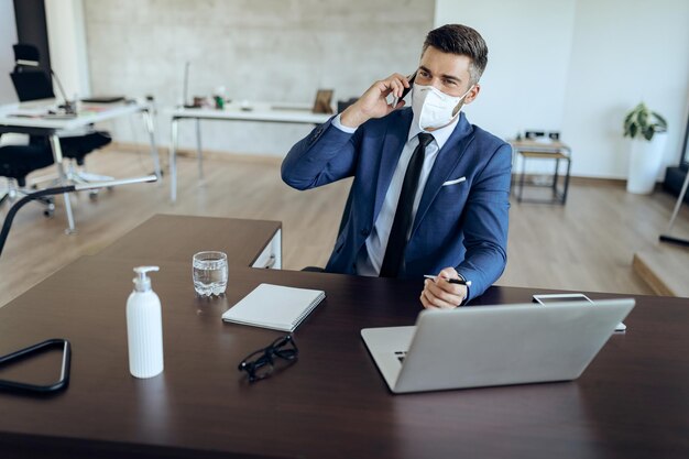 Zakenman die op mobiele telefoon praat terwijl hij een gezichtsmasker draagt en op kantoor werkt